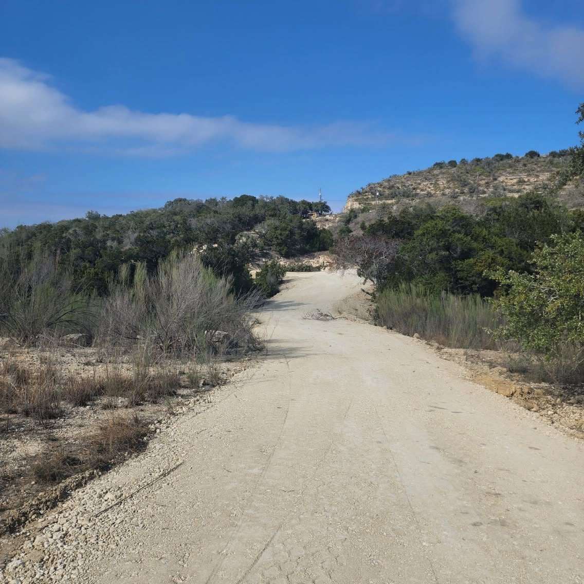 Wide graded road of pale crushed stone climbing a hillside through brush